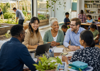 Adults collaborate around a table in a bright community classroom with children, bookshelves, and indoor garden beds